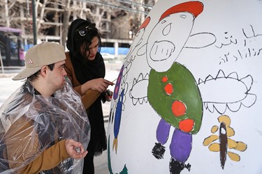 یکی از کودکان نقاش مبتلا به سندرم داون با کمک مربی خود در حال نقاشی بر روی تخم مرغ خود می باشد.