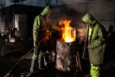 چند تن از پاکبان ها در محل سایت برف روبی در کنار آتش در شب برفی ایستاده اند