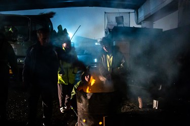 چند تن از پاکبان ها در محل سایت برف روبی در کنار آتش در شب برفی ایستاده اند