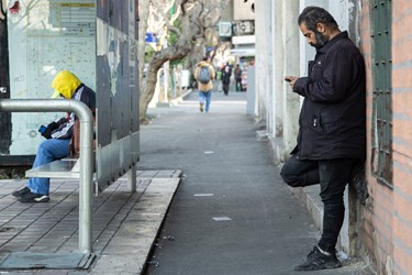 مردی  در انتظار آمدن اتوبوس در خیابان حافظ تهران است و  گوشی خود را چک میکند. 25 دی 1402