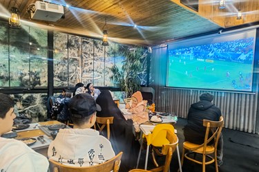 تعدادی از علاقمندان به فوتبال در یک کافه در کنار جمع خانوادگی  خود مشغول تماشای فوتبال ایران و قطر هستند